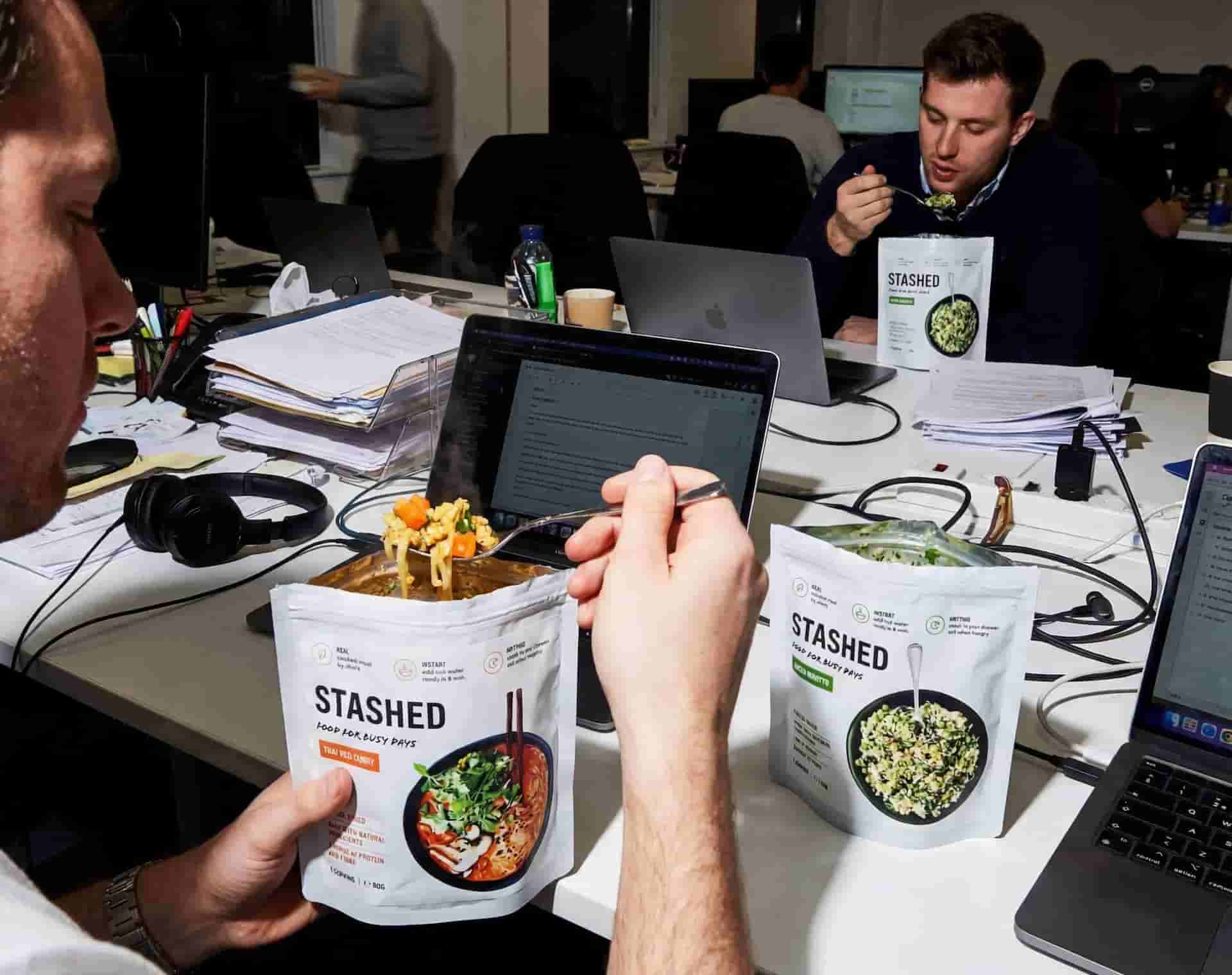 Person holding a 'Stashed' snack package in an office setting with colleagues working on laptops.